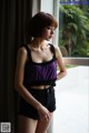 A woman standing by a window in a purple top and black shorts.